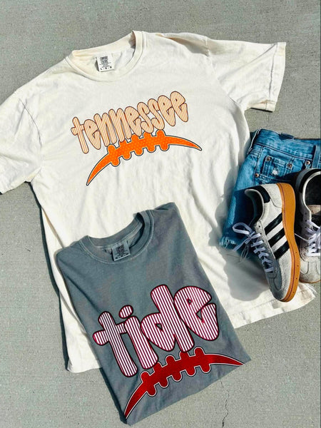 Two t-shirts with 'Tennessee' and 'tide' graphics on a gray surface with sneakers.