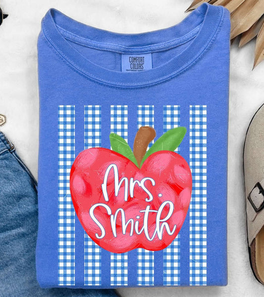 A folded blue T-shirt with a red apple graphic and the text 'Mrs. Smith' over blue-and-white gingham stripes, styled in a flat lay with jeans, sunglasses, a necklace, a slip-on shoe, and dried palm leaves on a white textured background.