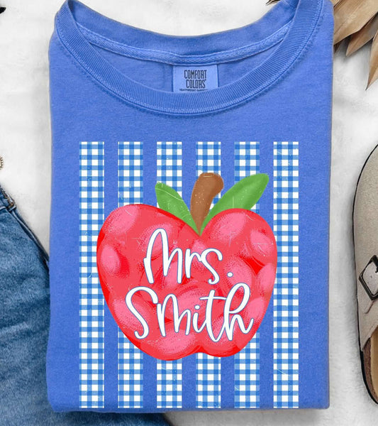 A folded blue T-shirt with a red apple graphic and the text 'Mrs. Smith' over blue-and-white gingham stripes, styled in a flat lay with jeans, sunglasses, a necklace, a slip-on shoe, and dried palm leaves on a white textured background.