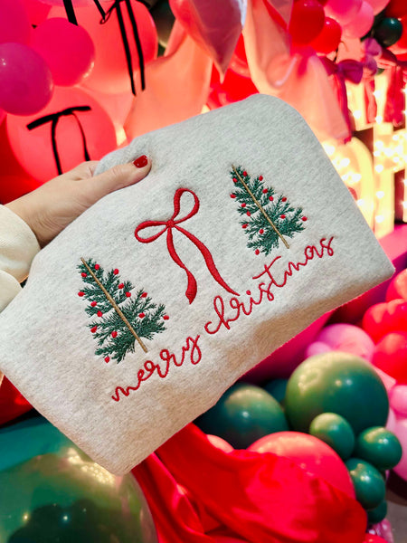 White sweatshirt folded with Christmas tree and bow design held by a hand against a colorful background of balloons.