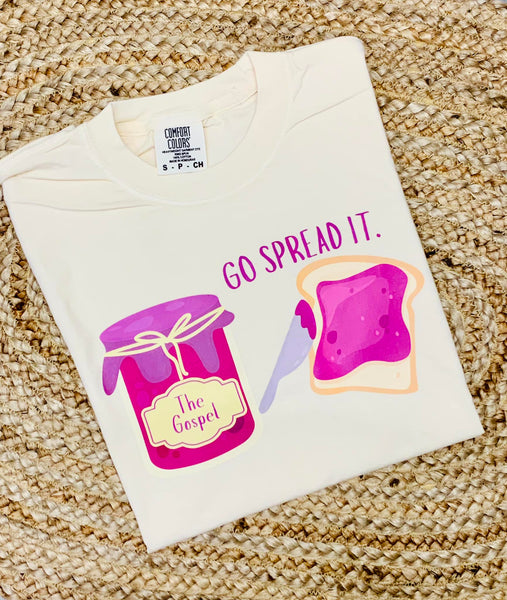 A youth-sized white t-shirt with a graphic print of a jar and a slice of bread, alongside the phrase 'GO SPREAD IT.'