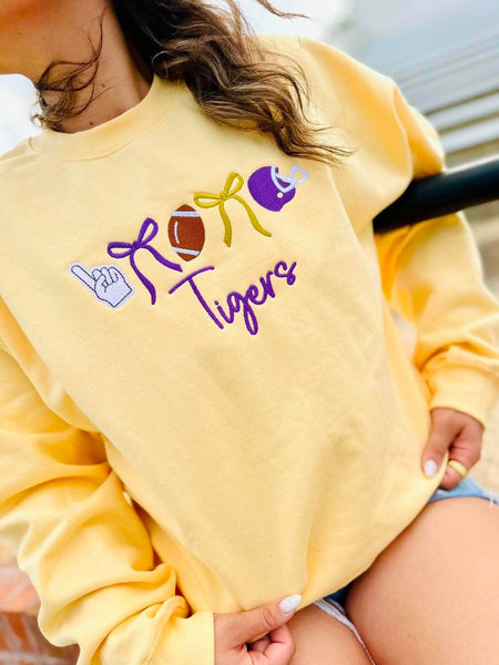 A person wearing a pale yellow sweatshirt with a 'ROAR Tigers' graphic featuring purple, yellow, and brown football-themed illustrations, paired with denim shorts.