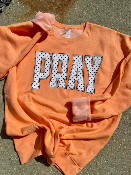 Orange sweatshirt with 'PRAY' printed on it in white letters with black polka dots, laid flat on a concrete surface.