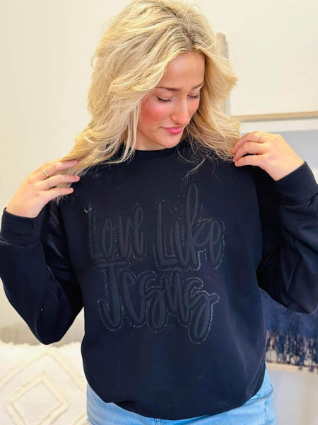 Woman wearing a black sweatshirt with Black monocromatic Love like Jesus , sitting on a couch.