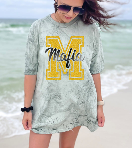 Woman wearing a gray dress with a yellow 'M' and the word 'Mafia' on a beach.