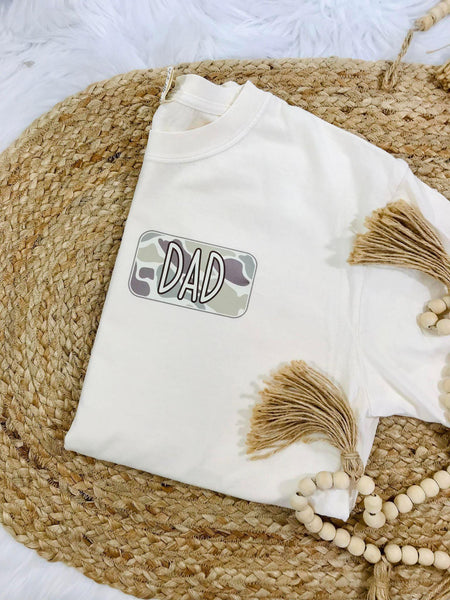 A white t-shirt with camouflage graphics and the text 'DAD' on the front pocket, displayed on a woven surface with beaded decorations in the background.