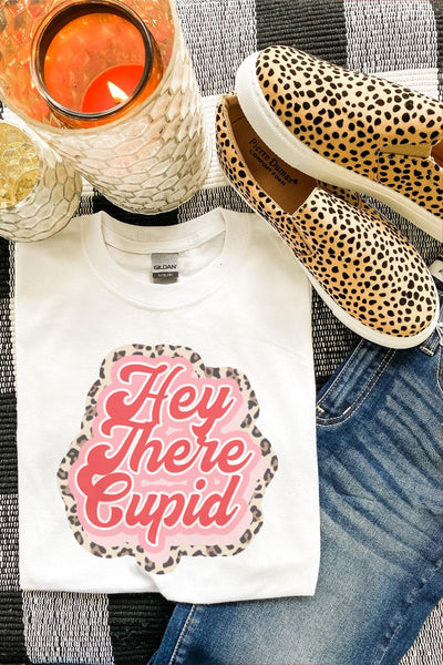 White t-shirt with 'Hey There Cupid' design, leopard print shoes, and candle on a checkered fabric background